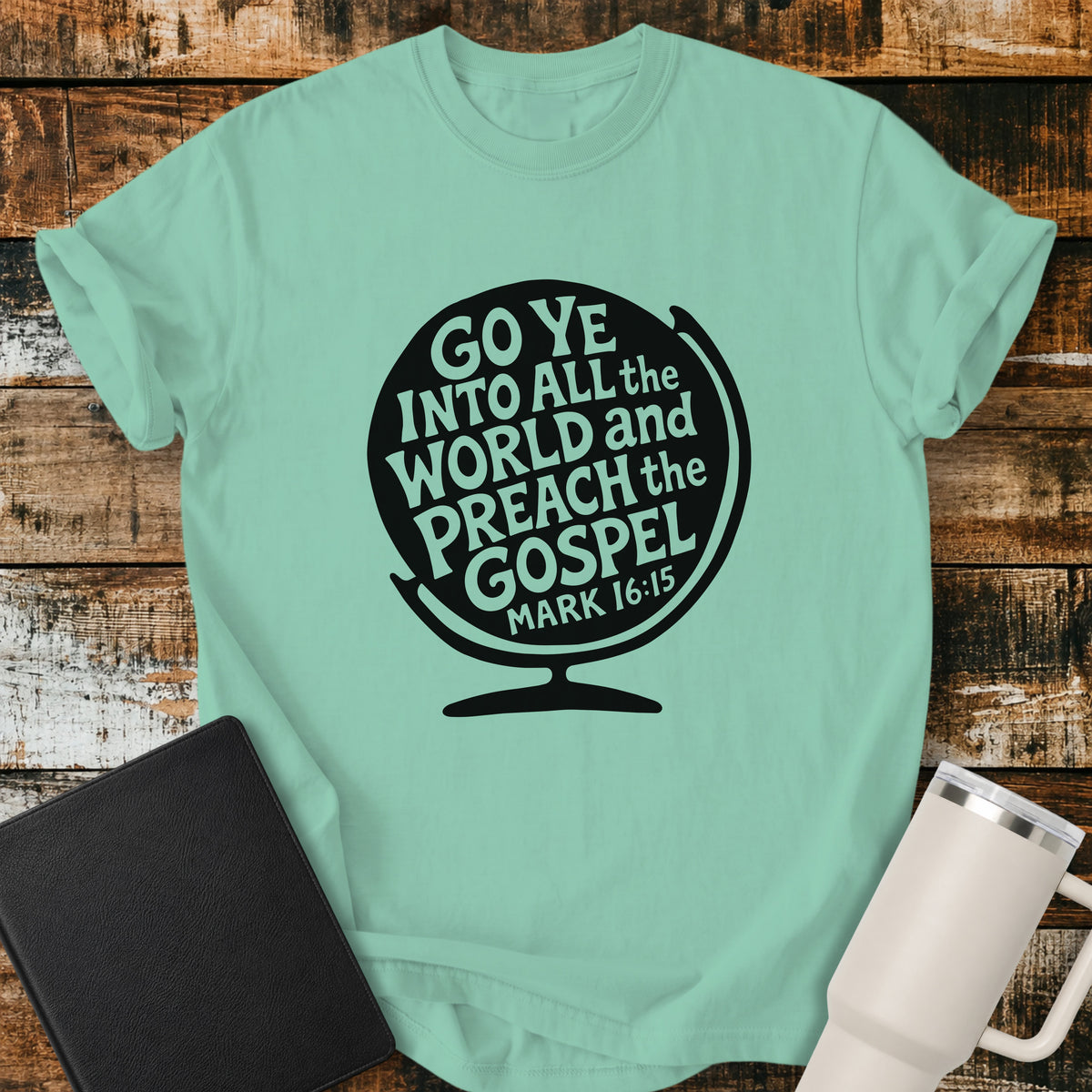 island reef green flat-lay t-shirt mockup featuring a black globe silhouette filled with stylized lettering that reads, "GO YE INTO ALL THE WORLD AND PREACH THE GOSPEL," with the Bible reference "MARK 16:15" at the bottom. The faith-inspired design is centered on the shirt and displayed on a rustic wood plank background alongside a black leather Bible and a white insulated travel mug.