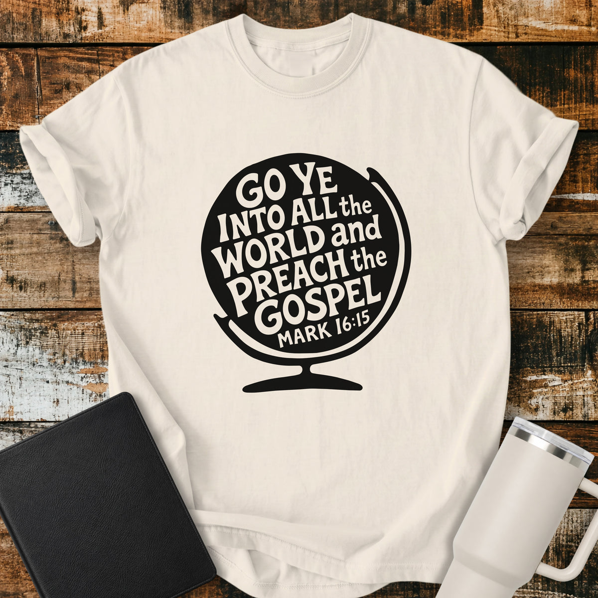 ivory flat-lay t-shirt mockup featuring a black globe silhouette filled with stylized lettering that reads, "GO YE INTO ALL THE WORLD AND PREACH THE GOSPEL," with the Bible reference "MARK 16:15" at the bottom. The faith-inspired design is centered on the shirt and displayed on a rustic wood plank background alongside a black leather Bible and a white insulated travel mug.