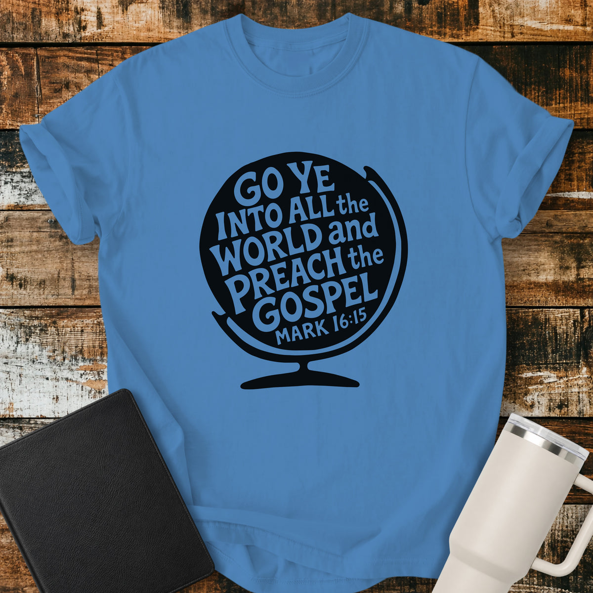 Royal caribe blue flat-lay t-shirt mockup featuring a black globe silhouette filled with stylized lettering that reads, "GO YE INTO ALL THE WORLD AND PREACH THE GOSPEL," with the Bible reference "MARK 16:15" at the bottom. The faith-inspired design is centered on the shirt and displayed on a rustic wood plank background alongside a black leather Bible and a white insulated travel mug.