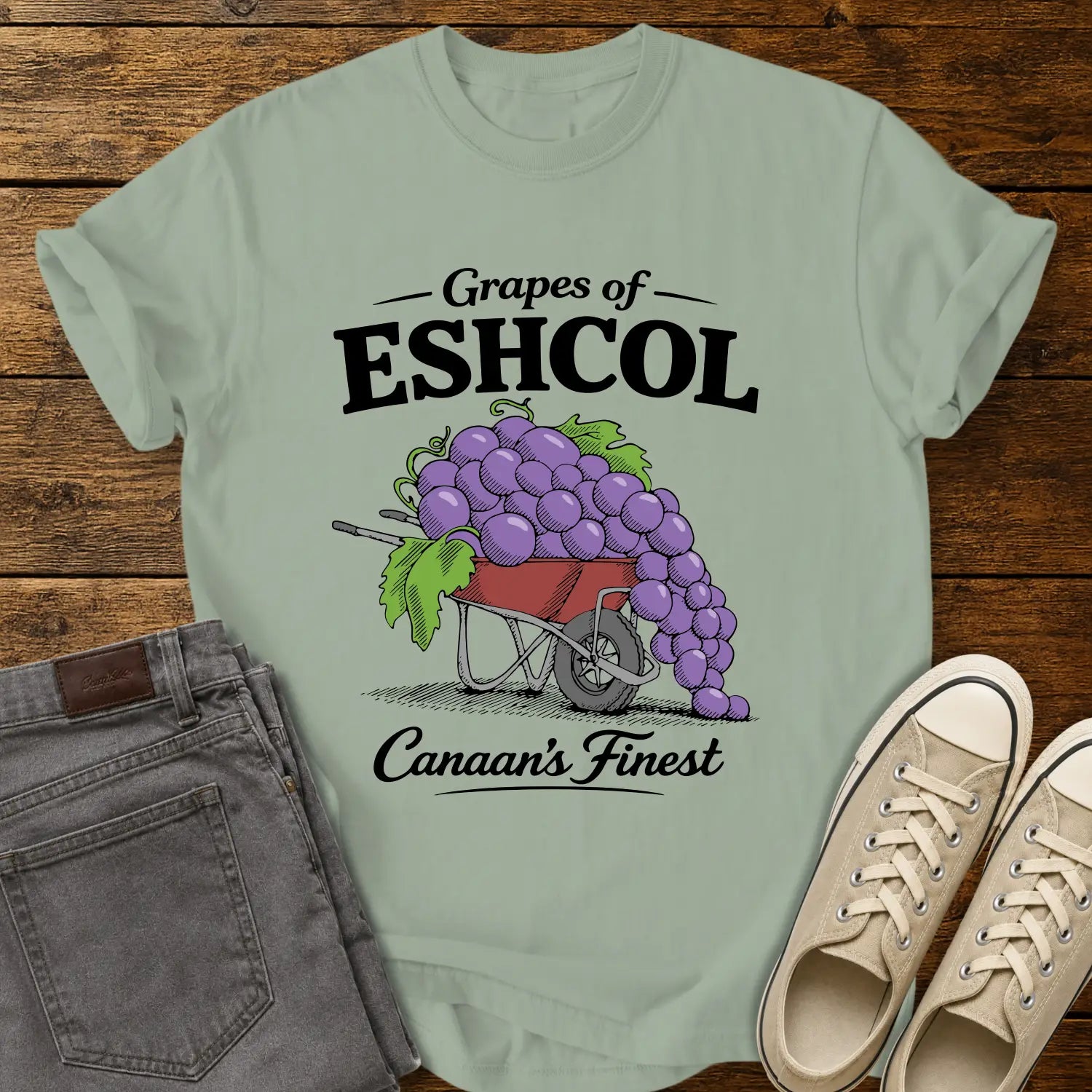 bay green graphic T-shirt mockup featuring a wheelbarrow overflowing with oversized purple grapes beneath the phrase “Grapes of Eshcol” and above the text “Canaan’s Finest,” styled with gray jeans and beige sneakers on a rustic wood background.