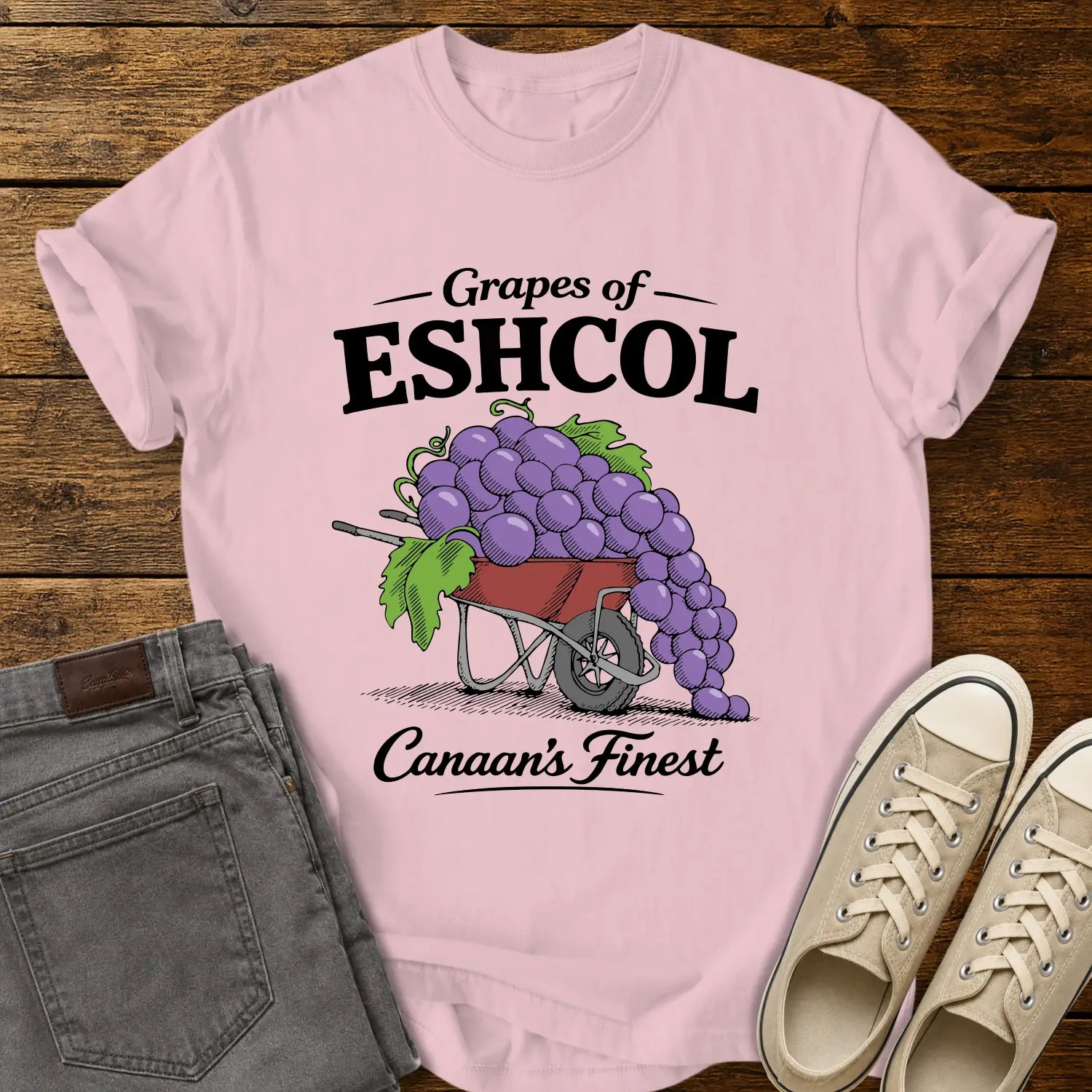 blossom pink graphic T-shirt mockup featuring a wheelbarrow overflowing with oversized purple grapes beneath the phrase “Grapes of Eshcol” and above the text “Canaan’s Finest,” styled with gray jeans and beige sneakers on a rustic wood background.
