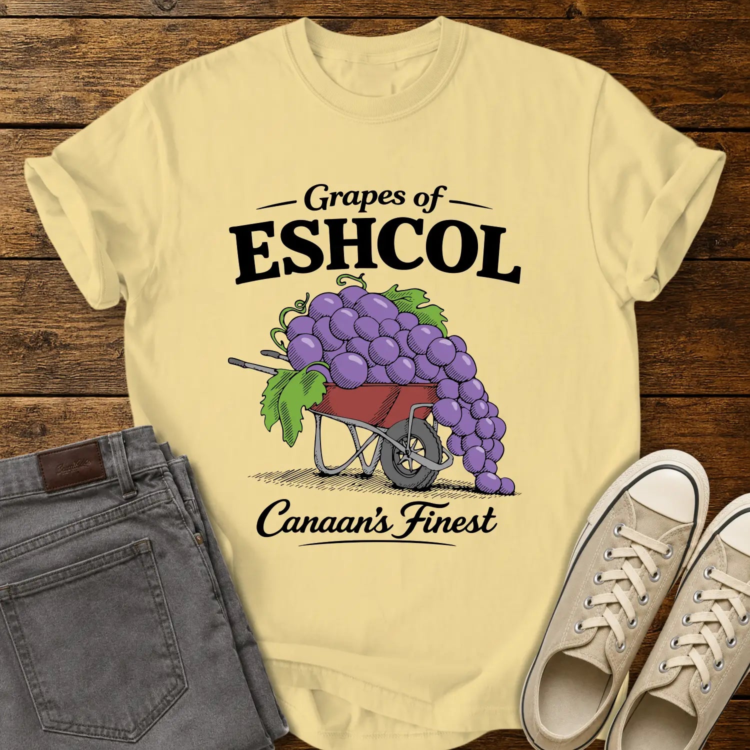 Butter yellow graphic T-shirt mockup featuring a wheelbarrow overflowing with oversized purple grapes beneath the phrase “Grapes of Eshcol” and above the text “Canaan’s Finest,” styled with gray jeans and beige sneakers on a rustic wood background.