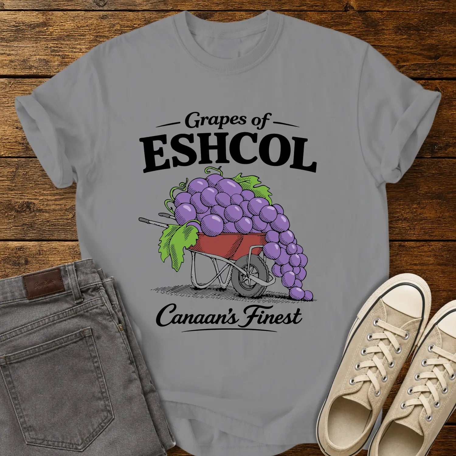 grey graphic T-shirt mockup featuring a wheelbarrow overflowing with oversized purple grapes beneath the phrase “Grapes of Eshcol” and above the text “Canaan’s Finest,” styled with gray jeans and beige sneakers on a rustic wood background.