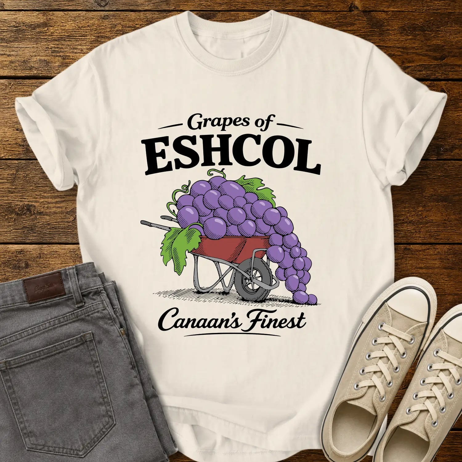 ivory graphic T-shirt mockup featuring a wheelbarrow overflowing with oversized purple grapes beneath the phrase “Grapes of Eshcol” and above the text “Canaan’s Finest,” styled with gray jeans and beige sneakers on a rustic wood background.
