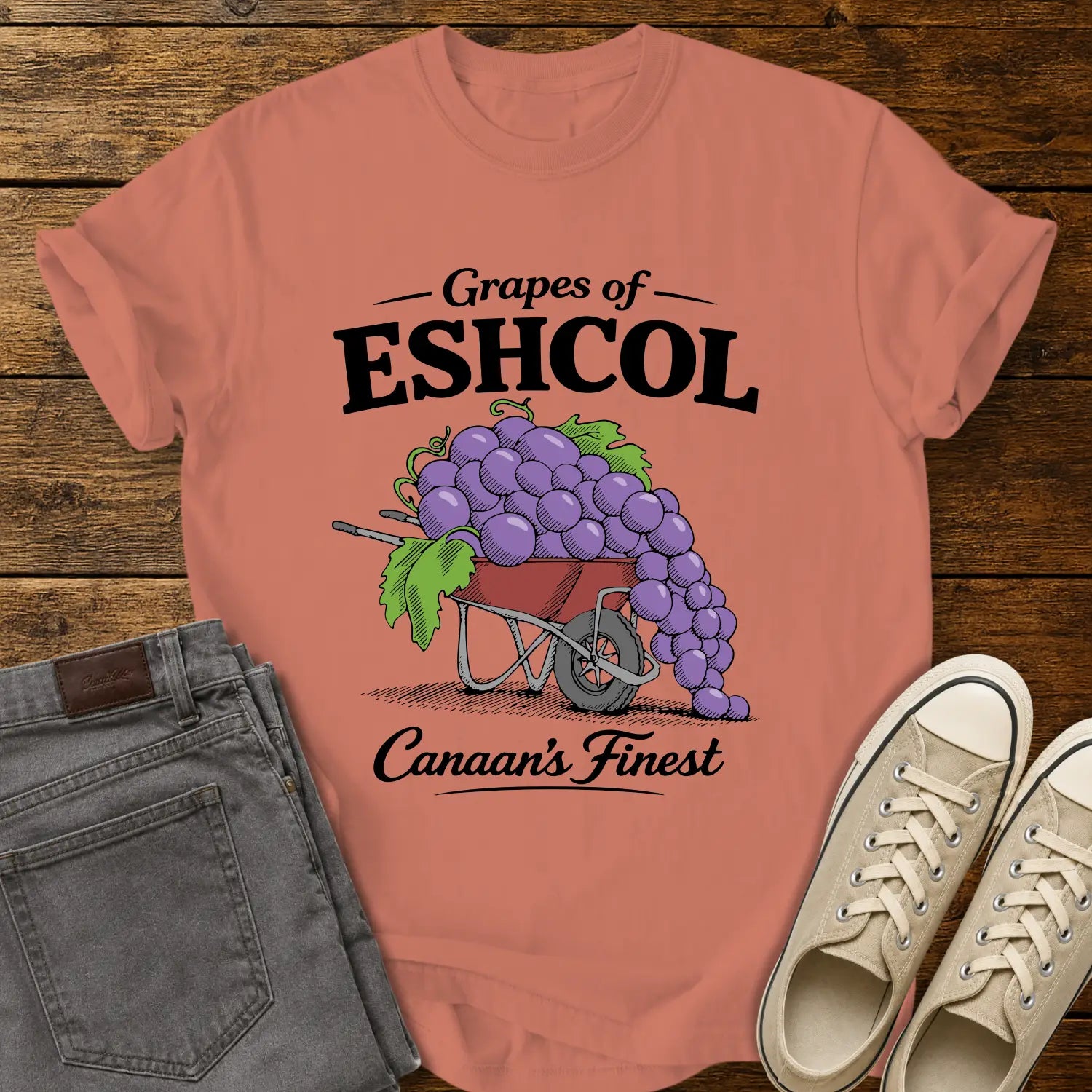 Terracotta graphic T-shirt mockup featuring a wheelbarrow overflowing with oversized purple grapes beneath the phrase “Grapes of Eshcol” and above the text “Canaan’s Finest,” styled with gray jeans and beige sneakers on a rustic wood background.