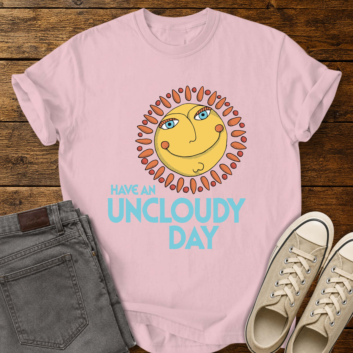 blossom pink flat-lay t-shirt mockup featuring a whimsical retro-style smiling sun with blue eyes and orange rays above the phrase, "HAVE AN UNCLOUDY DAY," in bold light blue lettering. The cheerful vintage-inspired design is centered on the shirt, which is displayed on a rustic wood plank background alongside folded gray jeans and a pair of cream canvas sneakers.