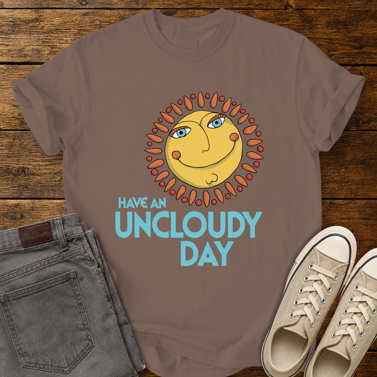 espresso flat-lay t-shirt mockup featuring a whimsical retro-style smiling sun with blue eyes and orange rays above the phrase, "HAVE AN UNCLOUDY DAY," in bold light blue lettering. The cheerful vintage-inspired design is centered on the shirt, which is displayed on a rustic wood plank background alongside folded gray jeans and a pair of cream canvas sneakers.
