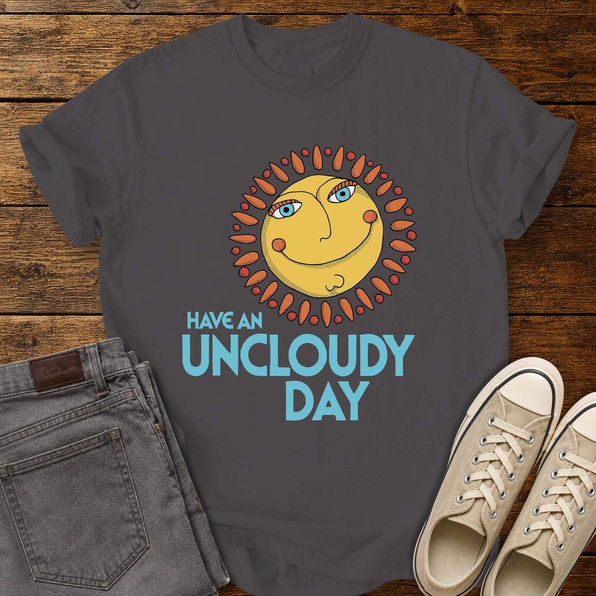 graphite flat-lay t-shirt mockup featuring a whimsical retro-style smiling sun with blue eyes and orange rays above the phrase, "HAVE AN UNCLOUDY DAY," in bold light blue lettering. The cheerful vintage-inspired design is centered on the shirt, which is displayed on a rustic wood plank background alongside folded gray jeans and a pair of cream canvas sneakers.