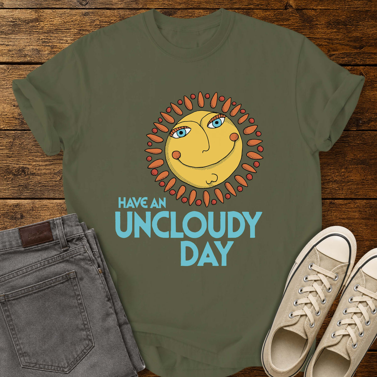 hemp green flat-lay t-shirt mockup featuring a whimsical retro-style smiling sun with blue eyes and orange rays above the phrase, "HAVE AN UNCLOUDY DAY," in bold light blue lettering. The cheerful vintage-inspired design is centered on the shirt, which is displayed on a rustic wood plank background alongside folded gray jeans and a pair of cream canvas sneakers.