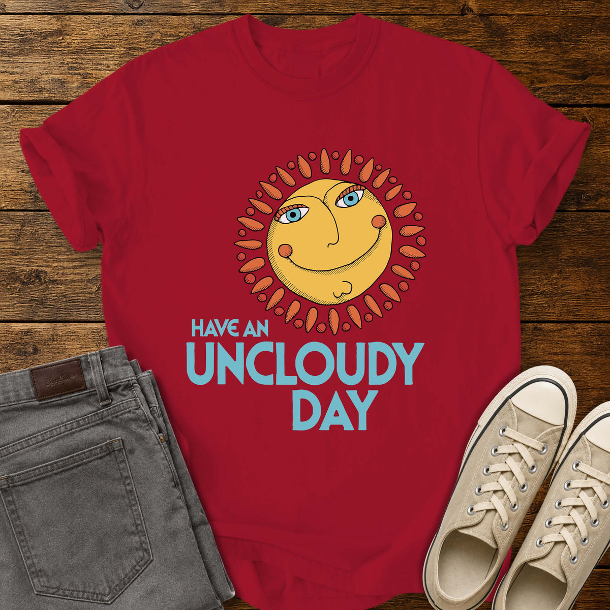 red flat-lay t-shirt mockup featuring a whimsical retro-style smiling sun with blue eyes and orange rays above the phrase, "HAVE AN UNCLOUDY DAY," in bold light blue lettering. The cheerful vintage-inspired design is centered on the shirt, which is displayed on a rustic wood plank background alongside folded gray jeans and a pair of cream canvas sneakers.