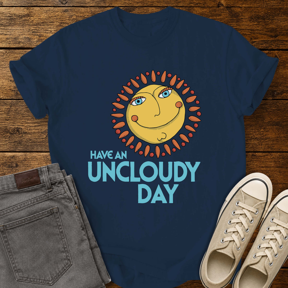 True navy flat-lay t-shirt mockup featuring a whimsical retro-style smiling sun with blue eyes and orange rays above the phrase, "HAVE AN UNCLOUDY DAY," in bold light blue lettering. The cheerful vintage-inspired design is centered on the shirt, which is displayed on a rustic wood plank background alongside folded gray jeans and a pair of cream canvas sneakers.