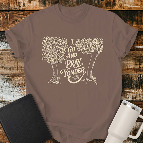 espresso T-shirt featuring a cream-colored vintage line-art illustration of two leafy trees framing the phrase, “I GO AND PRAY YONDER,” with “MATTHEW 26:36” beneath in smaller text. The design uses elegant retro lettering and a minimalist one-color print. The shirt is displayed on a rustic wood plank background with a black Bible on the left and a white insulated tumbler on the right. 
