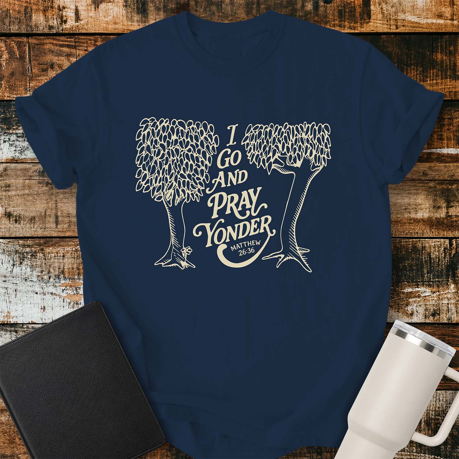 True navy T-shirt featuring a cream-colored vintage line-art illustration of two leafy trees framing the phrase, “I GO AND PRAY YONDER,” with “MATTHEW 26:36” beneath in smaller text. The design uses elegant retro lettering and a minimalist one-color print. The shirt is displayed on a rustic wood plank background with a black Bible on the left and a white insulated tumbler on the right.