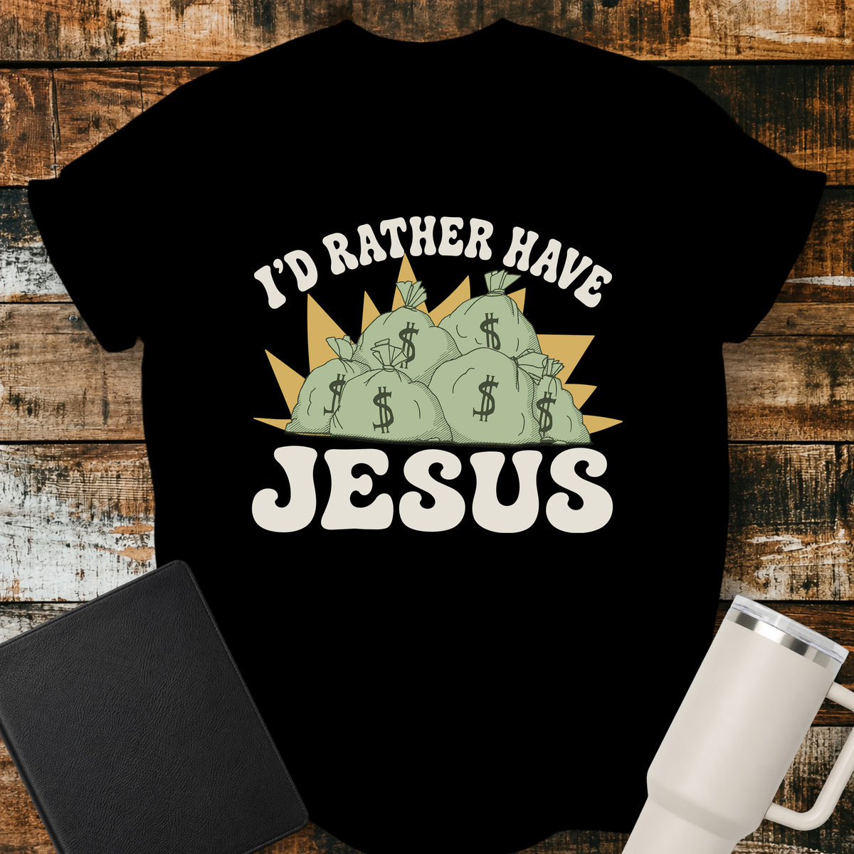 black flat-lay t-shirt mockup featuring a retro design with stacks of money bags beneath the phrase, "I'D RATHER HAVE," and the large word "JESUS" below. The vintage-inspired artwork contrasts earthly riches with faith using cream, green, and gold tones. The shirt is displayed on a rustic wood plank background alongside a black leather Bible and a white insulated travel mug.