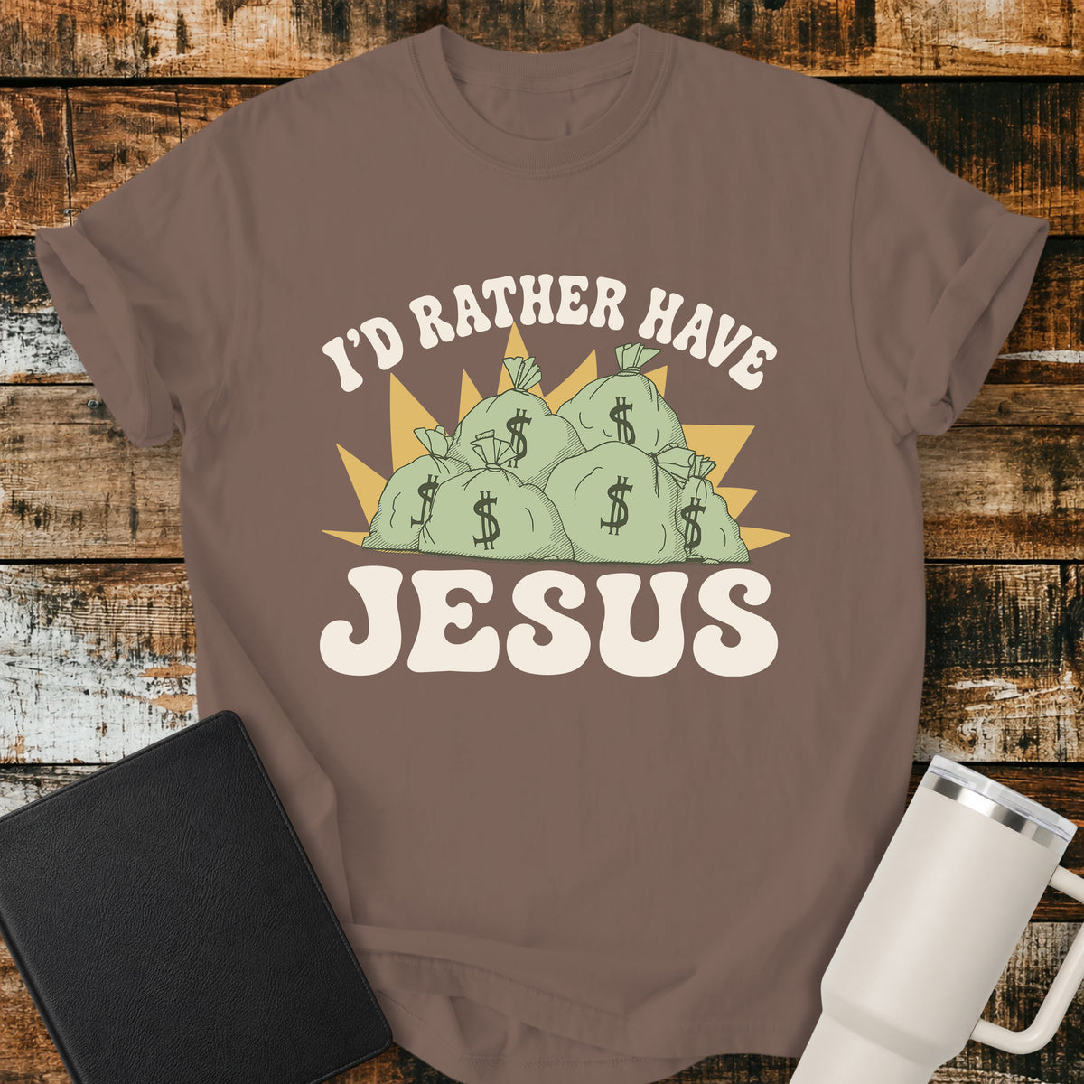 espresso brown flat-lay t-shirt mockup featuring a retro design with stacks of money bags beneath the phrase, "I'D RATHER HAVE," and the large word "JESUS" below. The vintage-inspired artwork contrasts earthly riches with faith using cream, green, and gold tones. The shirt is displayed on a rustic wood plank background alongside a black leather Bible and a white insulated travel mug.