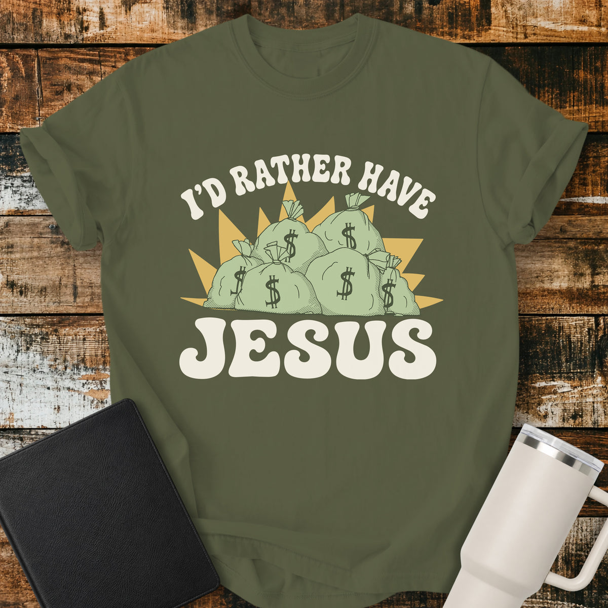 Hemp green flat-lay t-shirt mockup featuring a retro design with stacks of money bags beneath the phrase, "I'D RATHER HAVE," and the large word "JESUS" below. The vintage-inspired artwork contrasts earthly riches with faith using cream, green, and gold tones. The shirt is displayed on a rustic wood plank background alongside a black leather Bible and a white insulated travel mug.