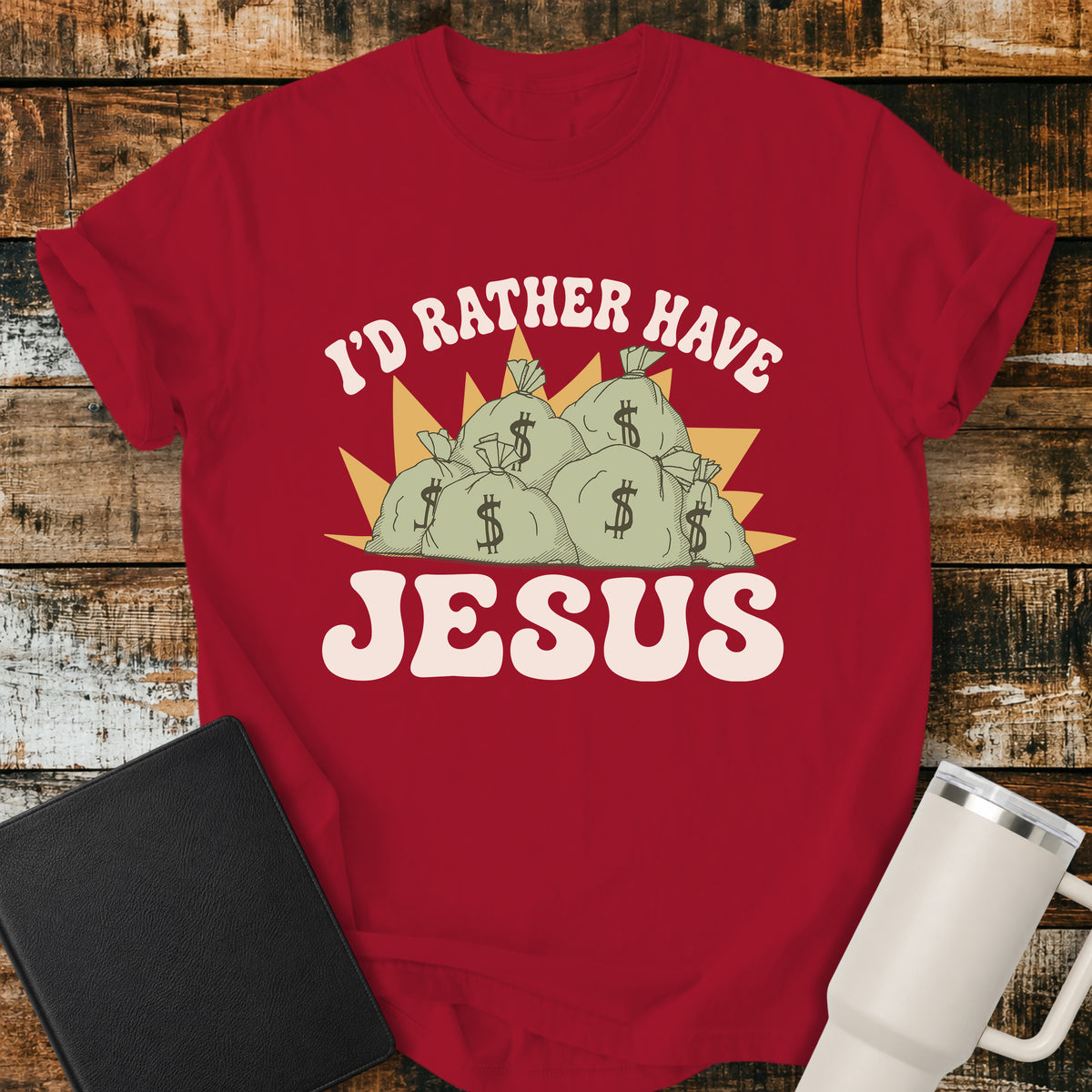 red flat-lay t-shirt mockup featuring a retro design with stacks of money bags beneath the phrase, "I'D RATHER HAVE," and the large word "JESUS" below. The vintage-inspired artwork contrasts earthly riches with faith using cream, green, and gold tones. The shirt is displayed on a rustic wood plank background alongside a black leather Bible and a white insulated travel mug.