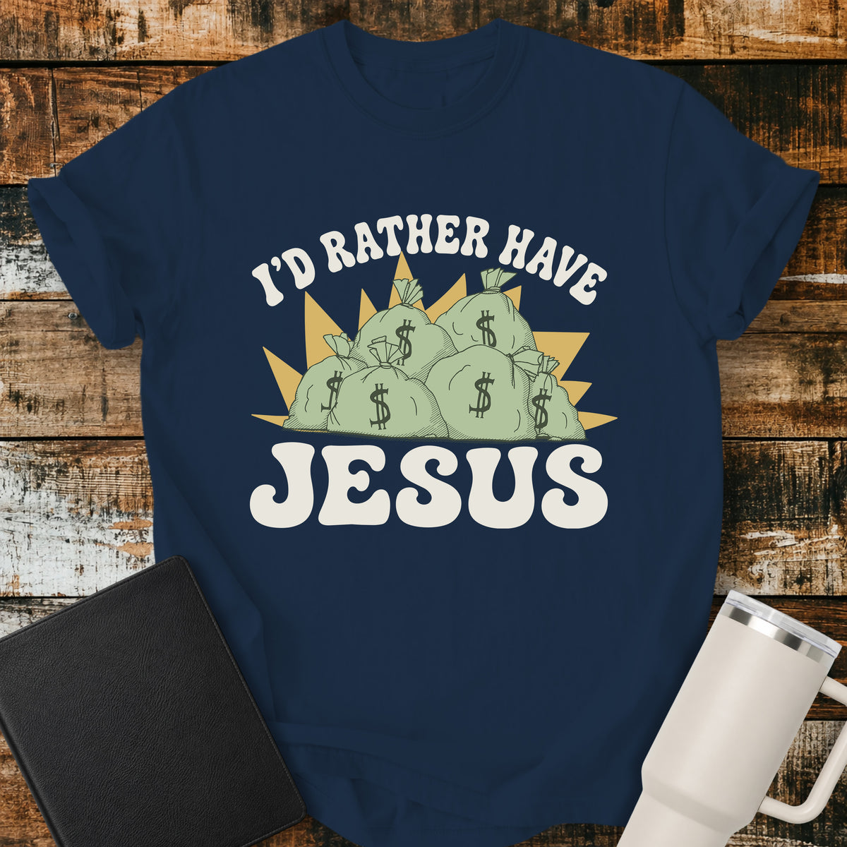 true navy flat-lay t-shirt mockup featuring a retro design with stacks of money bags beneath the phrase, "I'D RATHER HAVE," and the large word "JESUS" below. The vintage-inspired artwork contrasts earthly riches with faith using cream, green, and gold tones. The shirt is displayed on a rustic wood plank background alongside a black leather Bible and a white insulated travel mug.