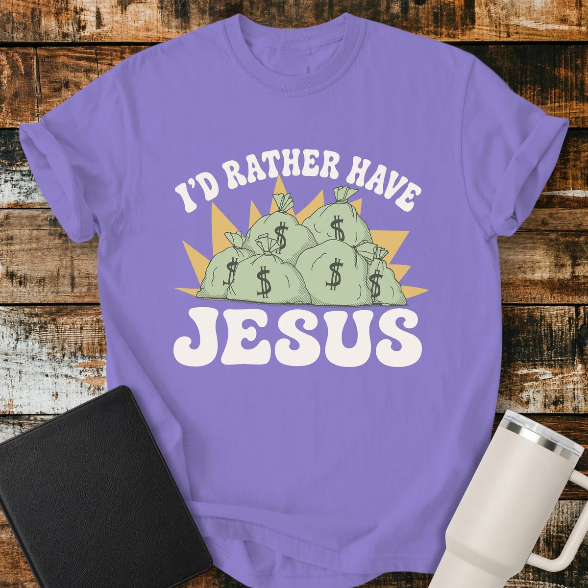 violet flat-lay t-shirt mockup featuring a retro design with stacks of money bags beneath the phrase, "I'D RATHER HAVE," and the large word "JESUS" below. The vintage-inspired artwork contrasts earthly riches with faith using cream, green, and gold tones. The shirt is displayed on a rustic wood plank background alongside a black leather Bible and a white insulated travel mug.