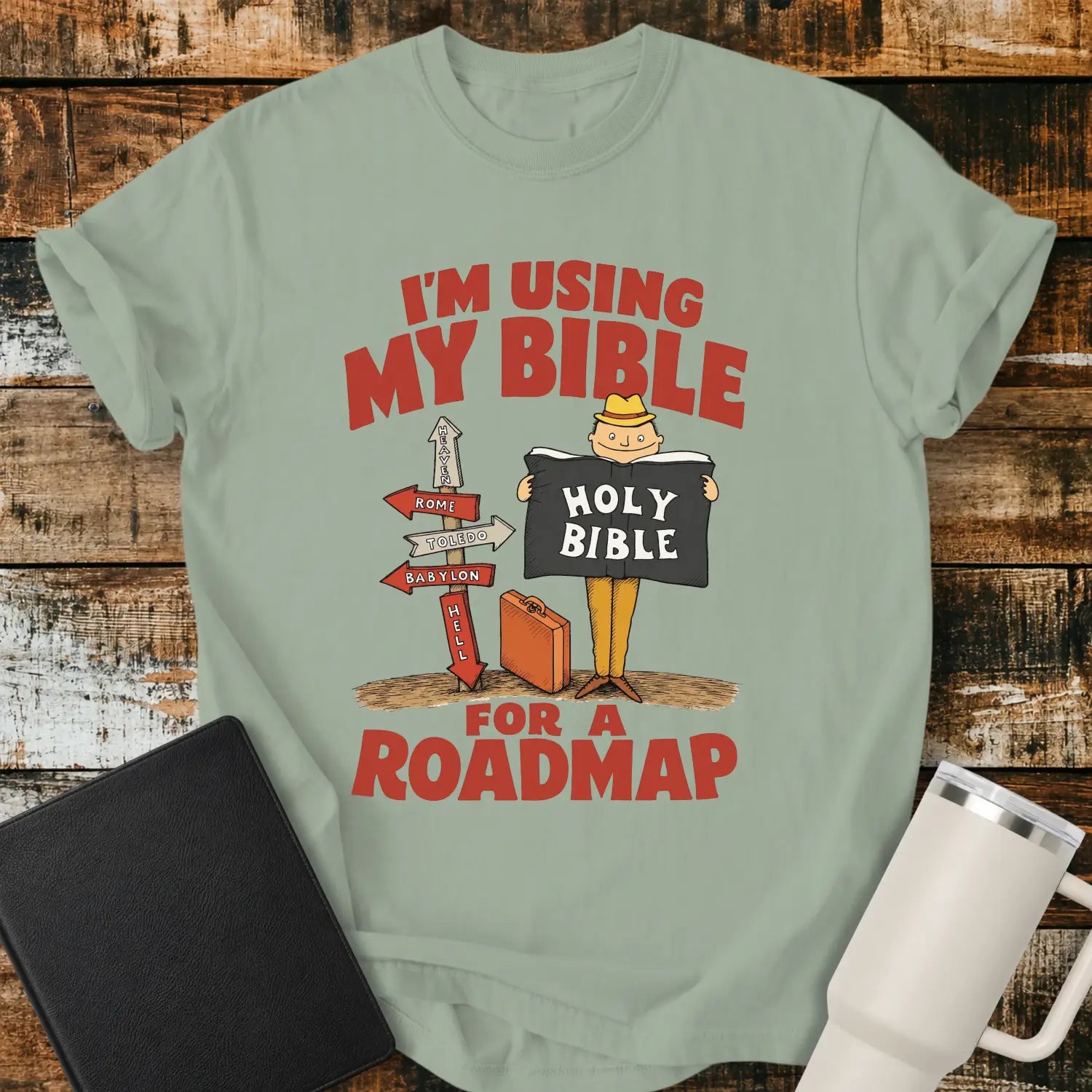 Bay green graphic T-shirt mockup featuring the phrase “I’m Using My Bible for a Roadmap” in bold red lettering with a cartoon traveler holding a Holy Bible beside directional signs, styled with a black Bible and white tumbler on a rustic wood background.