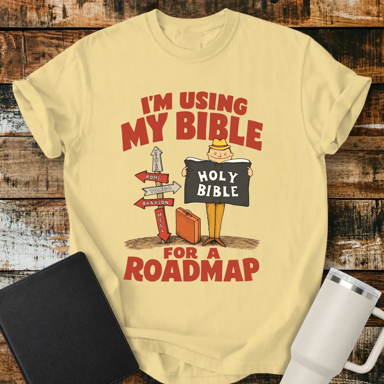 butter yellow graphic T-shirt mockup featuring the phrase “I’m Using My Bible for a Roadmap” in bold red lettering with a cartoon traveler holding a Holy Bible beside directional signs, styled with a black Bible and white tumbler on a rustic wood background.