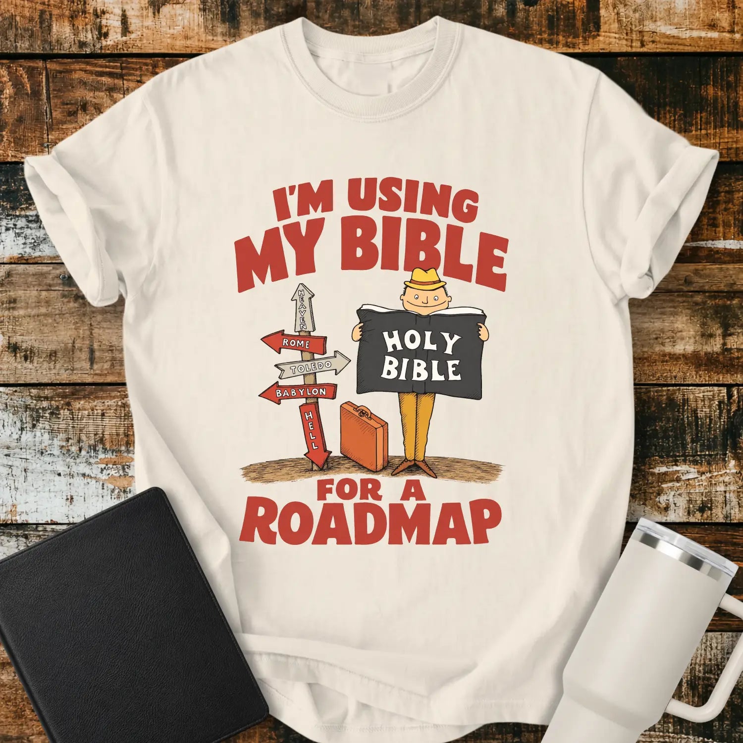 ivory graphic T-shirt mockup featuring the phrase “I’m Using My Bible for a Roadmap” in bold red lettering with a cartoon traveler holding a Holy Bible beside directional signs, styled with a black Bible and white tumbler on a rustic wood background.