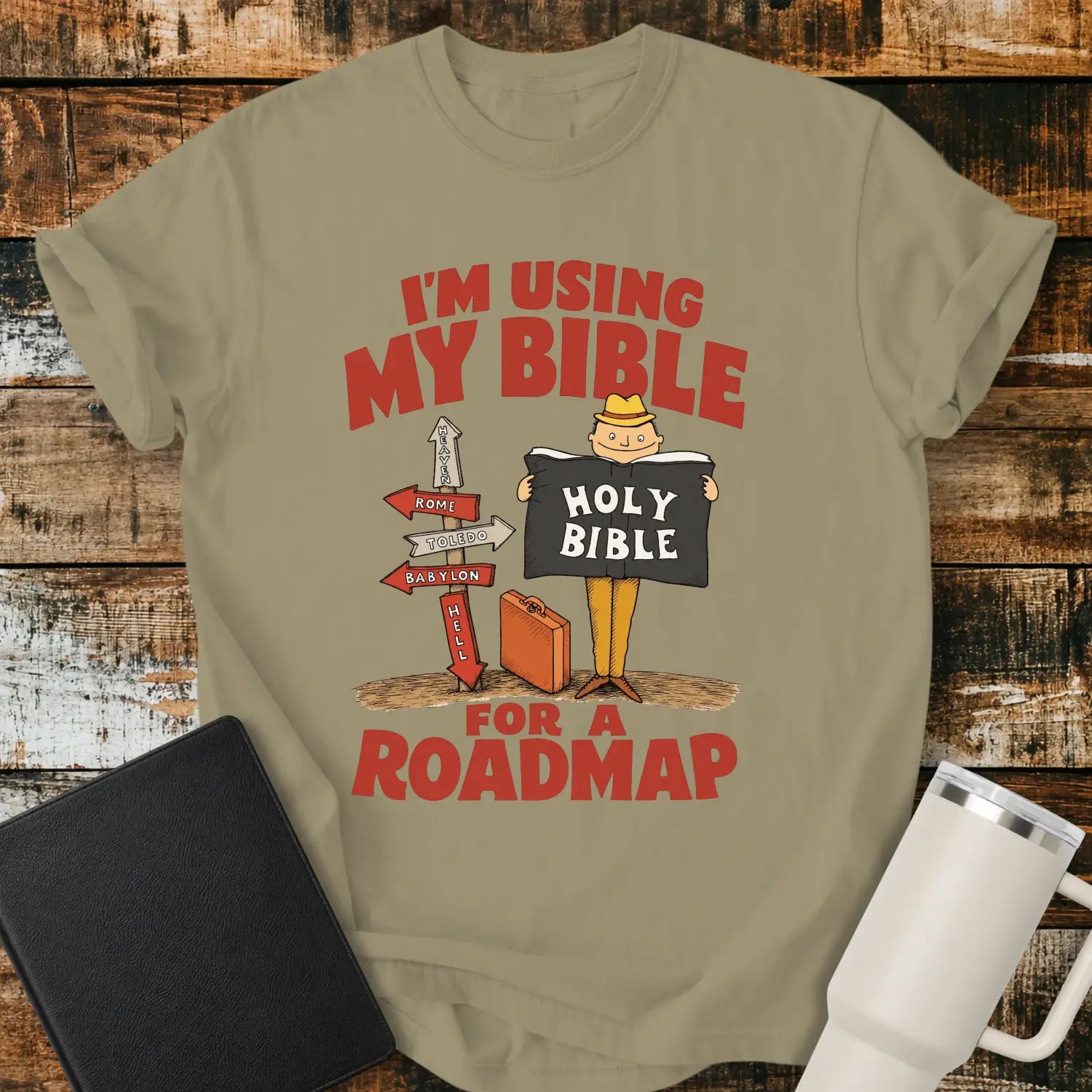 khaki graphic T-shirt mockup featuring the phrase “I’m Using My Bible for a Roadmap” in bold red lettering with a cartoon traveler holding a Holy Bible beside directional signs, styled with a black Bible and white tumbler on a rustic wood background.