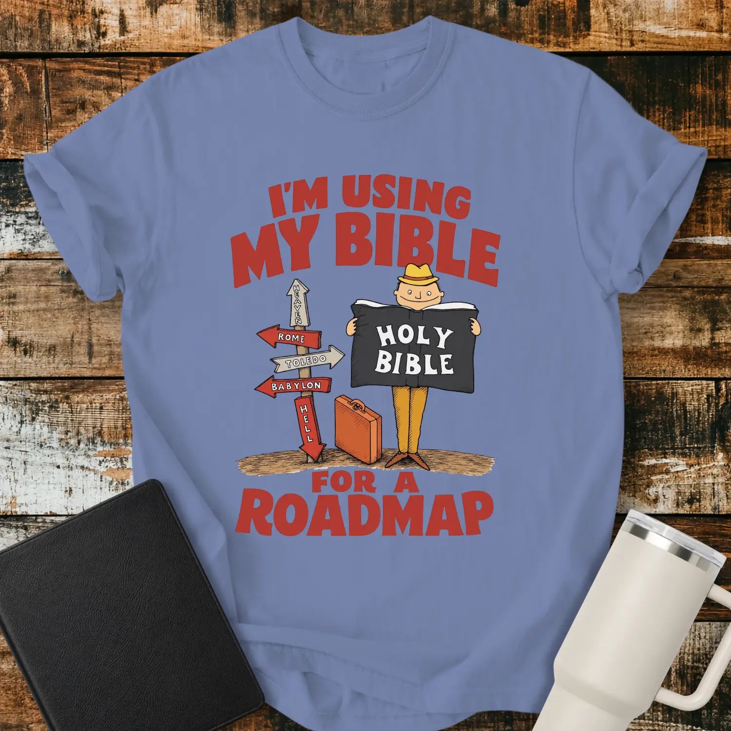 washed denim blue graphic T-shirt mockup featuring the phrase “I’m Using My Bible for a Roadmap” in bold red lettering with a cartoon traveler holding a Holy Bible beside directional signs, styled with a black Bible and white tumbler on a rustic wood background.
