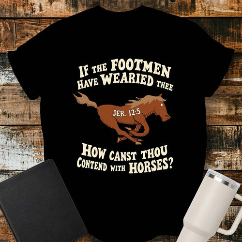black T-shirt featuring a running horse surrounded by the Bible verse, “IF THE FOOTMEN HAVE WEARIED THEE, HOW CANST THOU CONTEND WITH HORSES?” with “JER. 12:5” on the horse. The shirt is displayed on a rustic wood plank background alongside a black Bible and a white insulated tumbler.