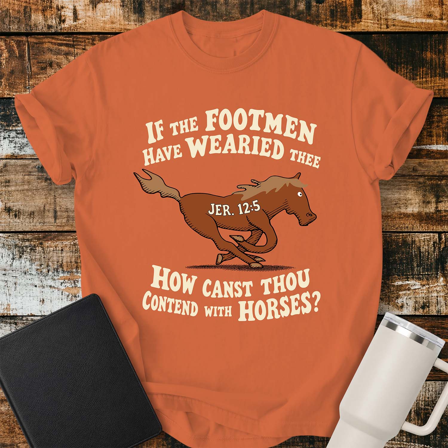 Burnt orange T-shirt featuring a running horse surrounded by the Bible verse, “IF THE FOOTMEN HAVE WEARIED THEE, HOW CANST THOU CONTEND WITH HORSES?” with “JER. 12:5” on the horse. The shirt is displayed on a rustic wood plank background alongside a black Bible and a white insulated tumbler.