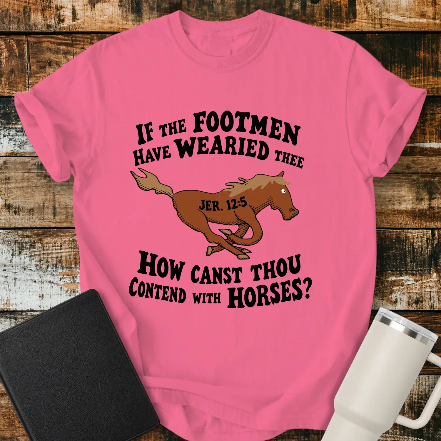 crunchberry pink graphic T-shirt mockup featuring a running horse illustration with the Bible verse phrase “If the Footmen Have Wearied Thee, How Canst Thou Contend with Horses?” and Jeremiah 12:5 reference, styled with a black Bible and white tumbler on a rustic wood background.