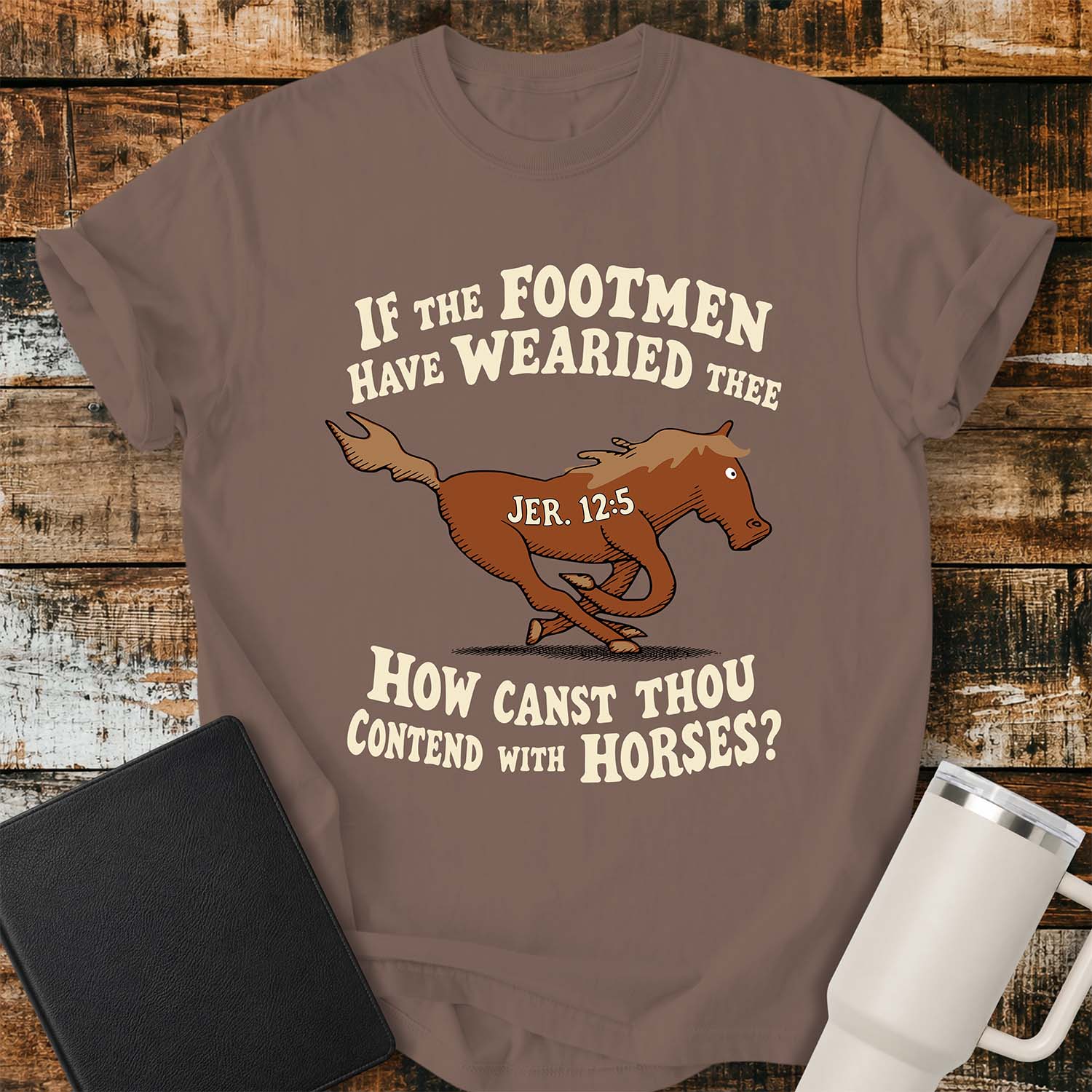 espresso T-shirt featuring a running horse surrounded by the Bible verse, “IF THE FOOTMEN HAVE WEARIED THEE, HOW CANST THOU CONTEND WITH HORSES?” with “JER. 12:5” on the horse. The shirt is displayed on a rustic wood plank background alongside a black Bible and a white insulated tumbler.