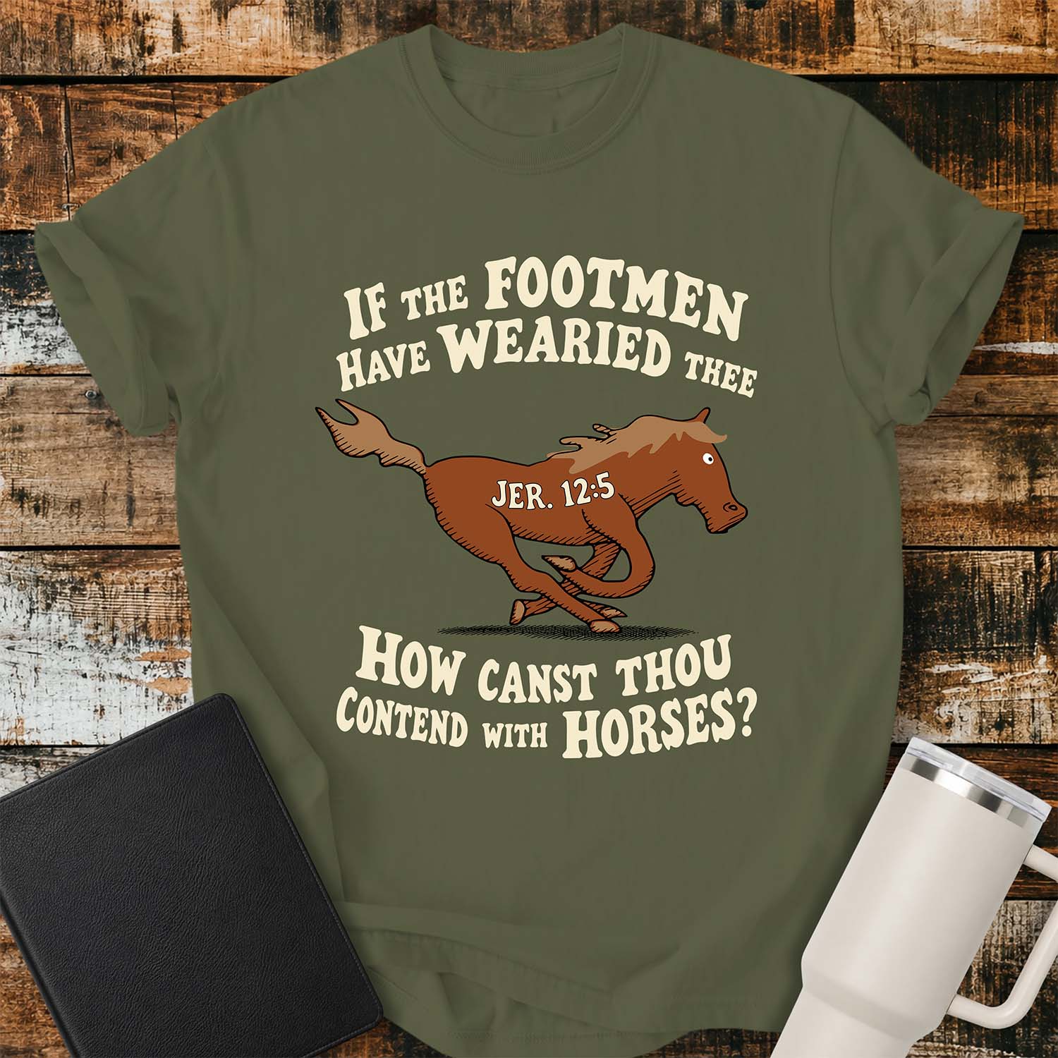 hemp green T-shirt featuring a running horse surrounded by the Bible verse, “IF THE FOOTMEN HAVE WEARIED THEE, HOW CANST THOU CONTEND WITH HORSES?” with “JER. 12:5” on the horse. The shirt is displayed on a rustic wood plank background alongside a black Bible and a white insulated tumbler.