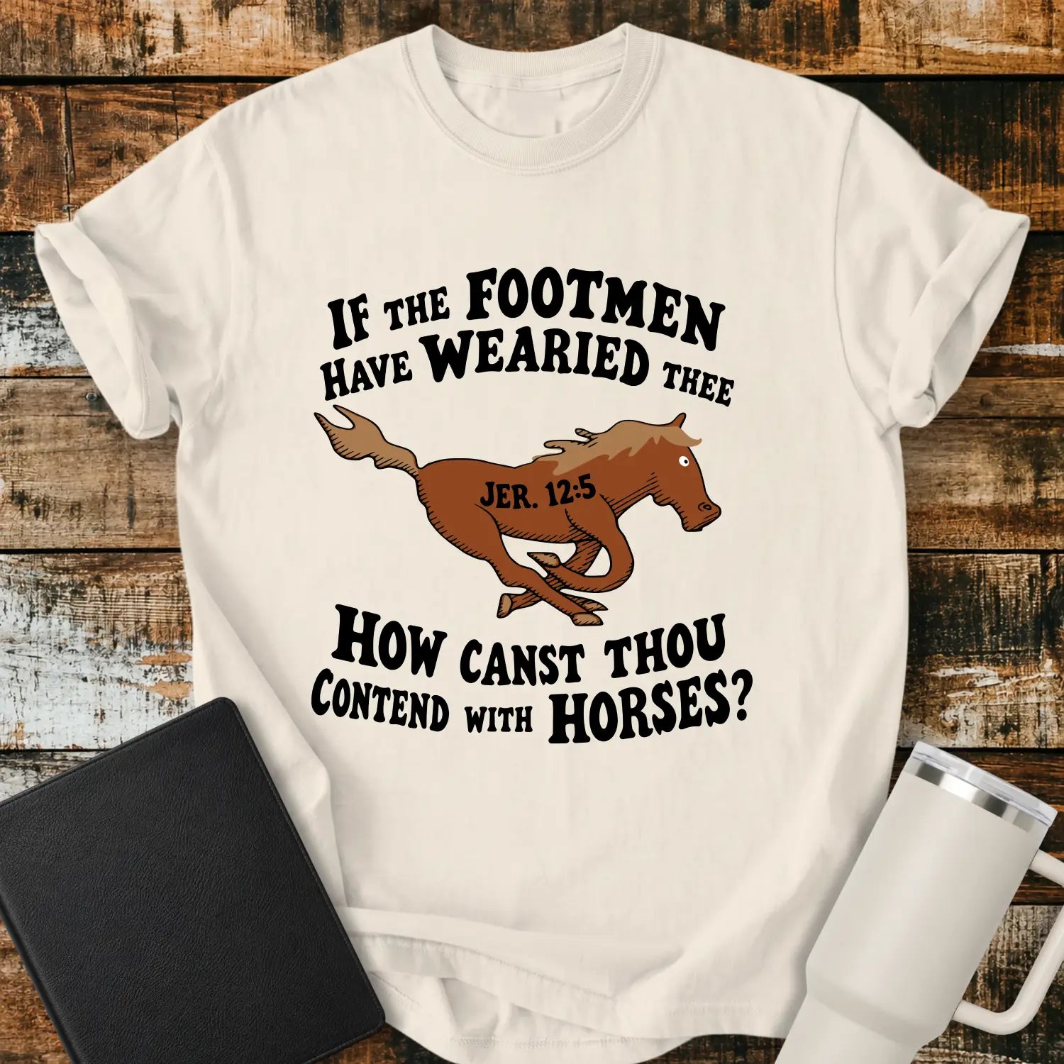 ivory graphic T-shirt mockup featuring a running horse illustration with the Bible verse phrase “If the Footmen Have Wearied Thee, How Canst Thou Contend with Horses?” and Jeremiah 12:5 reference, styled with a black Bible and white tumbler on a rustic wood background.