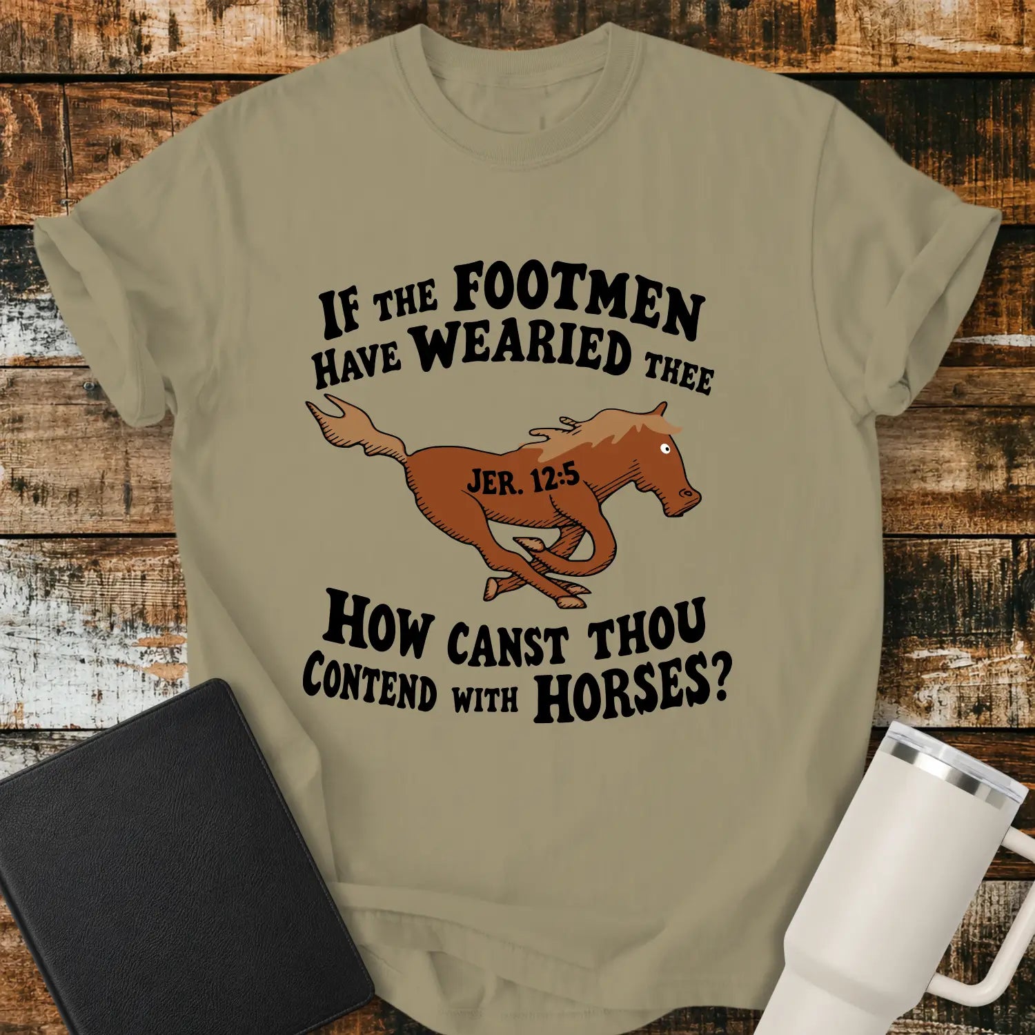 khaki graphic T-shirt mockup featuring a running horse illustration with the Bible verse phrase “If the Footmen Have Wearied Thee, How Canst Thou Contend with Horses?” and Jeremiah 12:5 reference, styled with a black Bible and white tumbler on a rustic wood background.