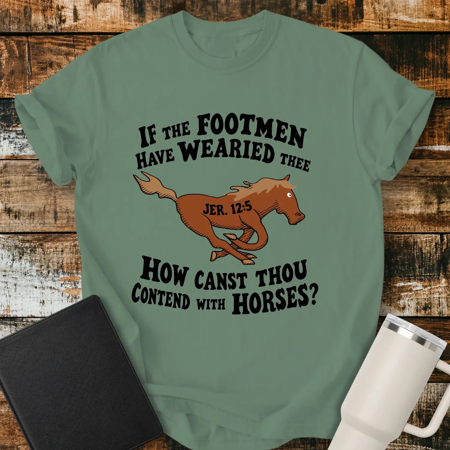 light green graphic T-shirt mockup featuring a running horse illustration with the Bible verse phrase “If the Footmen Have Wearied Thee, How Canst Thou Contend with Horses?” and Jeremiah 12:5 reference, styled with a black Bible and white tumbler on a rustic wood background.