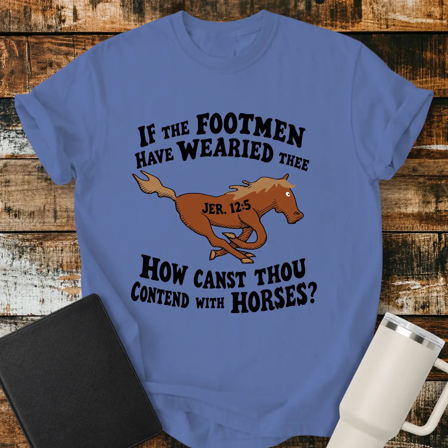 Mystic blue graphic T-shirt mockup featuring a running horse illustration with the Bible verse phrase “If the Footmen Have Wearied Thee, How Canst Thou Contend with Horses?” and Jeremiah 12:5 reference, styled with a black Bible and white tumbler on a rustic wood background.