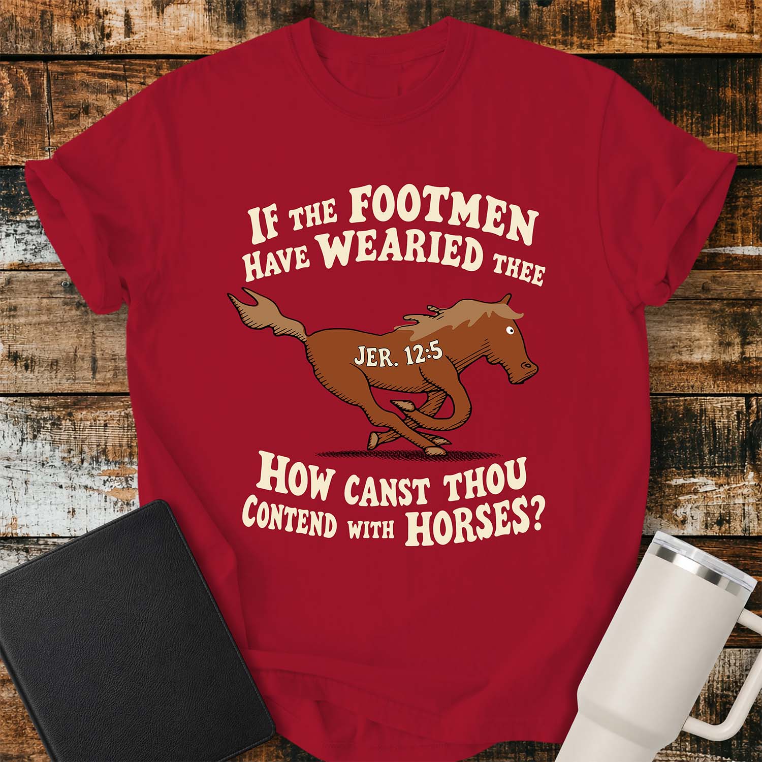 red T-shirt featuring a running horse surrounded by the Bible verse, “IF THE FOOTMEN HAVE WEARIED THEE, HOW CANST THOU CONTEND WITH HORSES?” with “JER. 12:5” on the horse. The shirt is displayed on a rustic wood plank background alongside a black Bible and a white insulated tumbler.