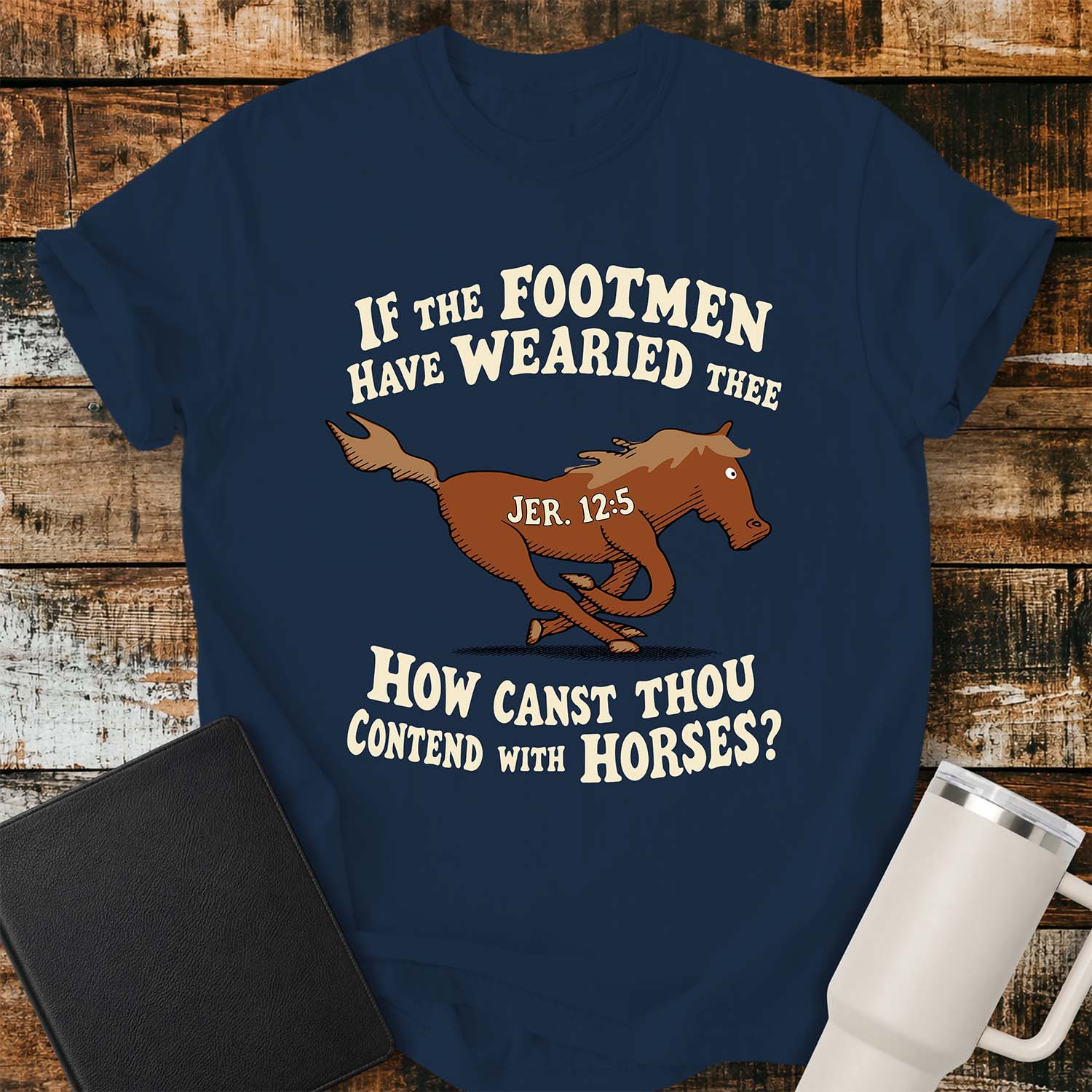true-navy-T-shirt featuring a running horse surrounded by the Bible verse, “IF THE FOOTMEN HAVE WEARIED THEE, HOW CANST THOU CONTEND WITH HORSES?” with “JER. 12:5” on the horse. The shirt is displayed on a rustic wood plank background alongside a black Bible and a white insulated tumbler.