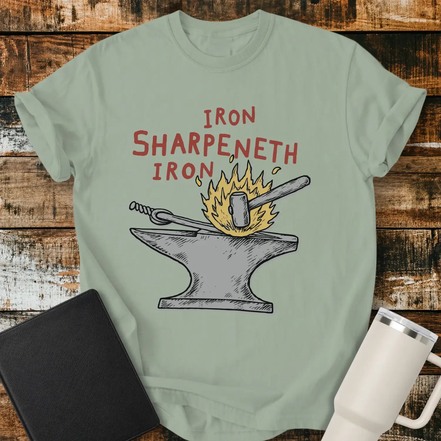 bay green graphic T-shirt mockup featuring the phrase “Iron Sharpeneth Iron” in red lettering above an illustration of a blacksmith’s anvil, hammer, heated iron rod, and sparks, styled with a black Bible and white tumbler on a rustic wood background.