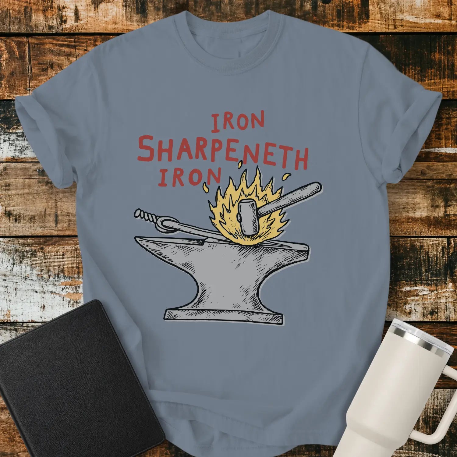 blue jean graphic T-shirt mockup featuring the phrase “Iron Sharpeneth Iron” in red lettering above an illustration of a blacksmith’s anvil, hammer, heated iron rod, and sparks, styled with a black Bible and white tumbler on a rustic wood background.