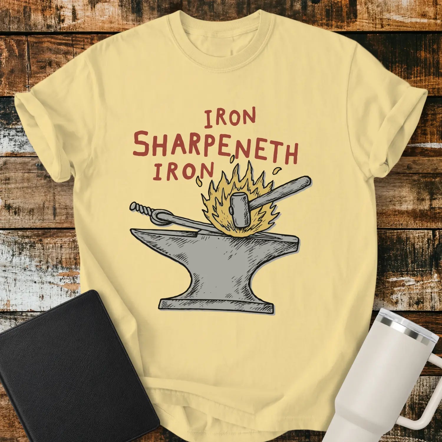 butter graphic T-shirt mockup featuring the phrase “Iron Sharpeneth Iron” in red lettering above an illustration of a blacksmith’s anvil, hammer, heated iron rod, and sparks, styled with a black Bible and white tumbler on a rustic wood background.