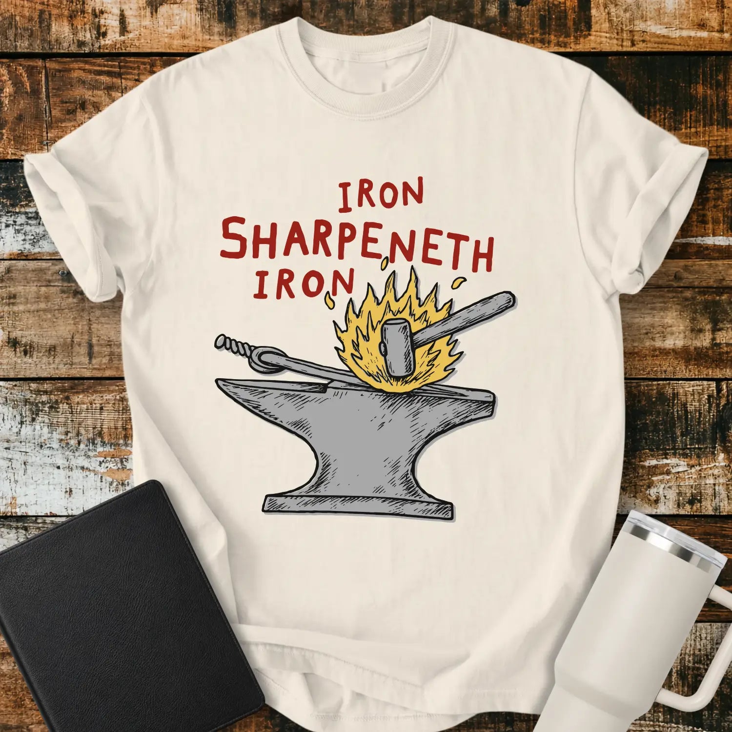 ivory graphic T-shirt mockup featuring the phrase “Iron Sharpeneth Iron” in red lettering above an illustration of a blacksmith’s anvil, hammer, heated iron rod, and sparks, styled with a black Bible and white tumbler on a rustic wood background.