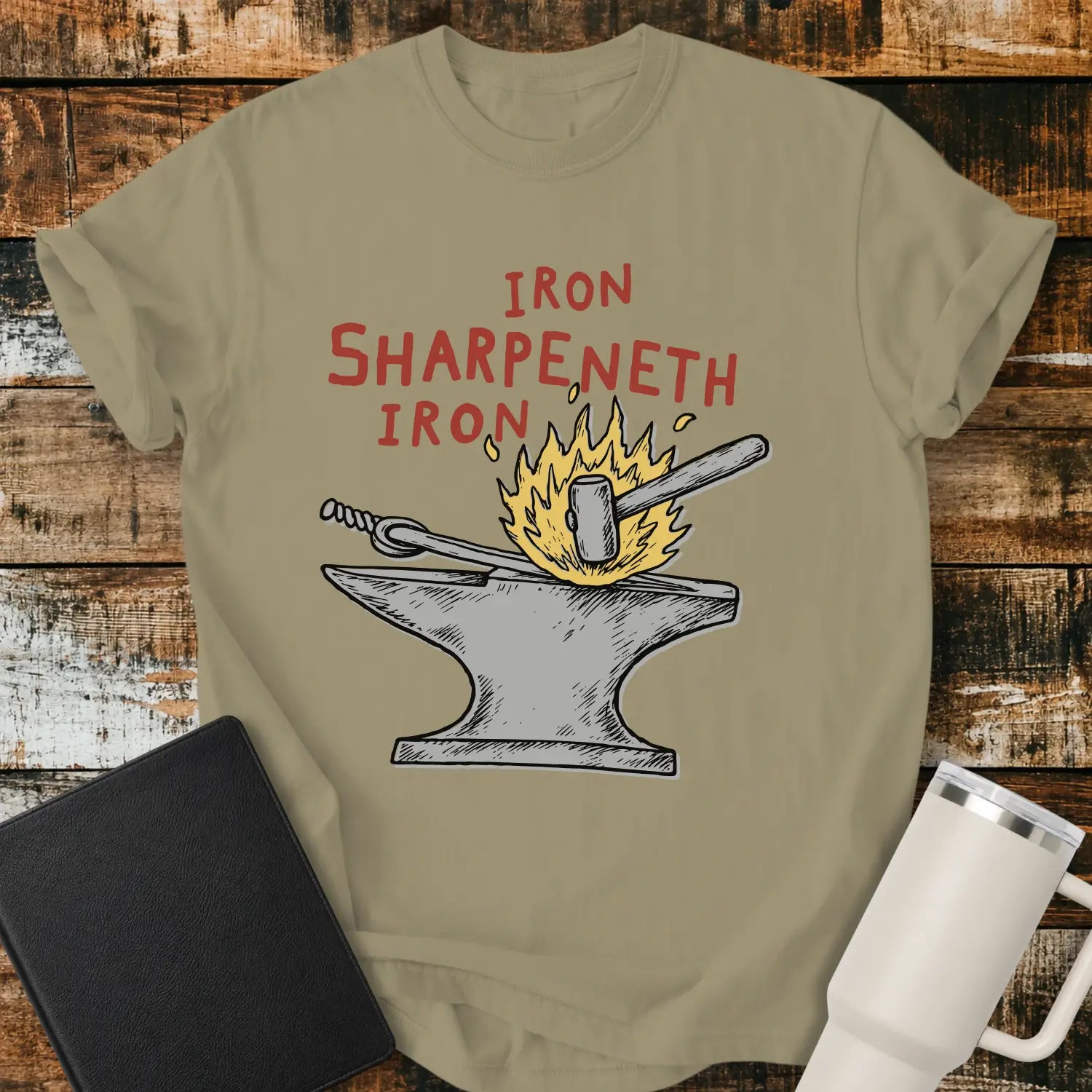 Khaki graphic T-shirt mockup featuring the phrase “Iron Sharpeneth Iron” in red lettering above an illustration of a blacksmith’s anvil, hammer, heated iron rod, and sparks, styled with a black Bible and white tumbler on a rustic wood background.