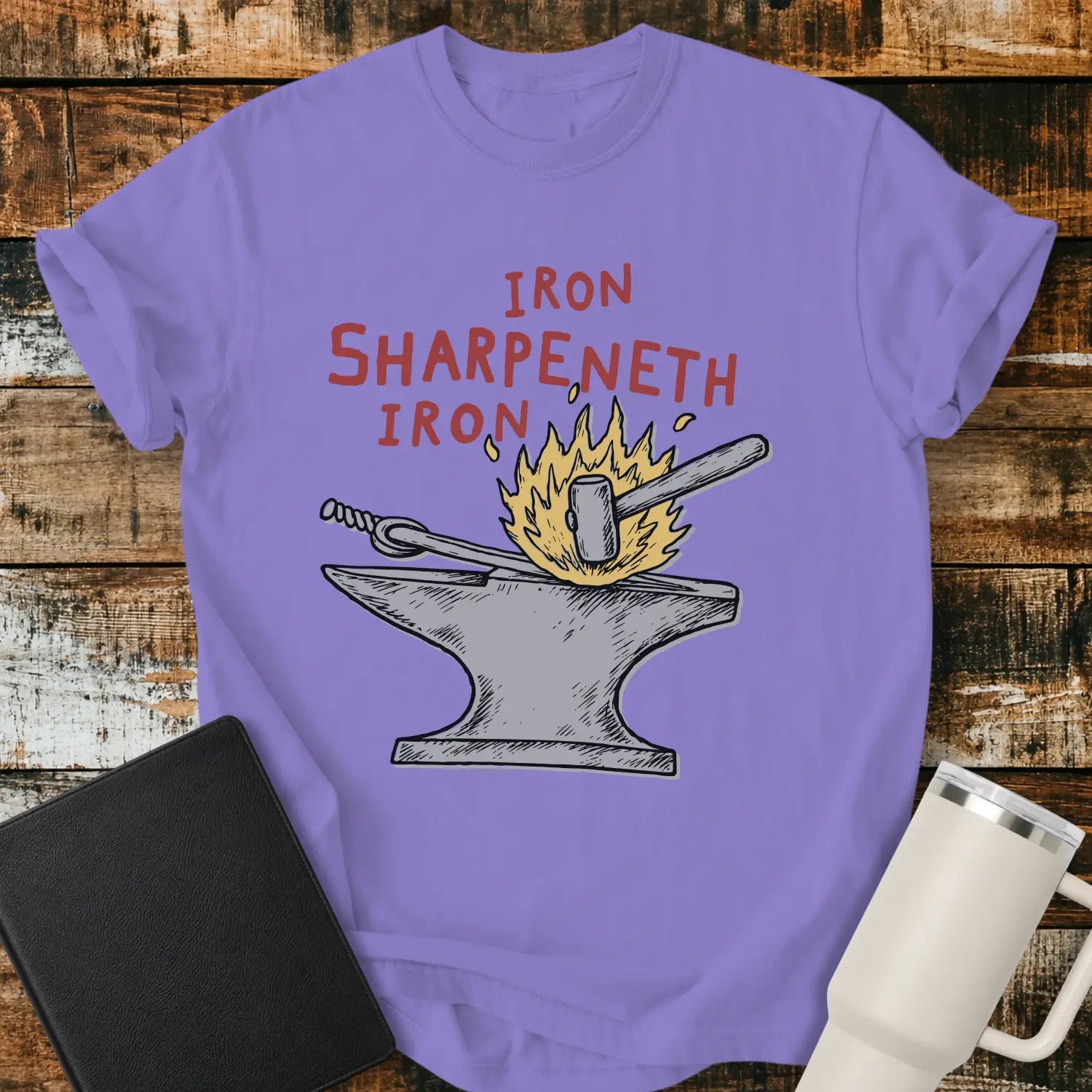 violet graphic T-shirt mockup featuring the phrase “Iron Sharpeneth Iron” in red lettering above an illustration of a blacksmith’s anvil, hammer, heated iron rod, and sparks, styled with a black Bible and white tumbler on a rustic wood background.