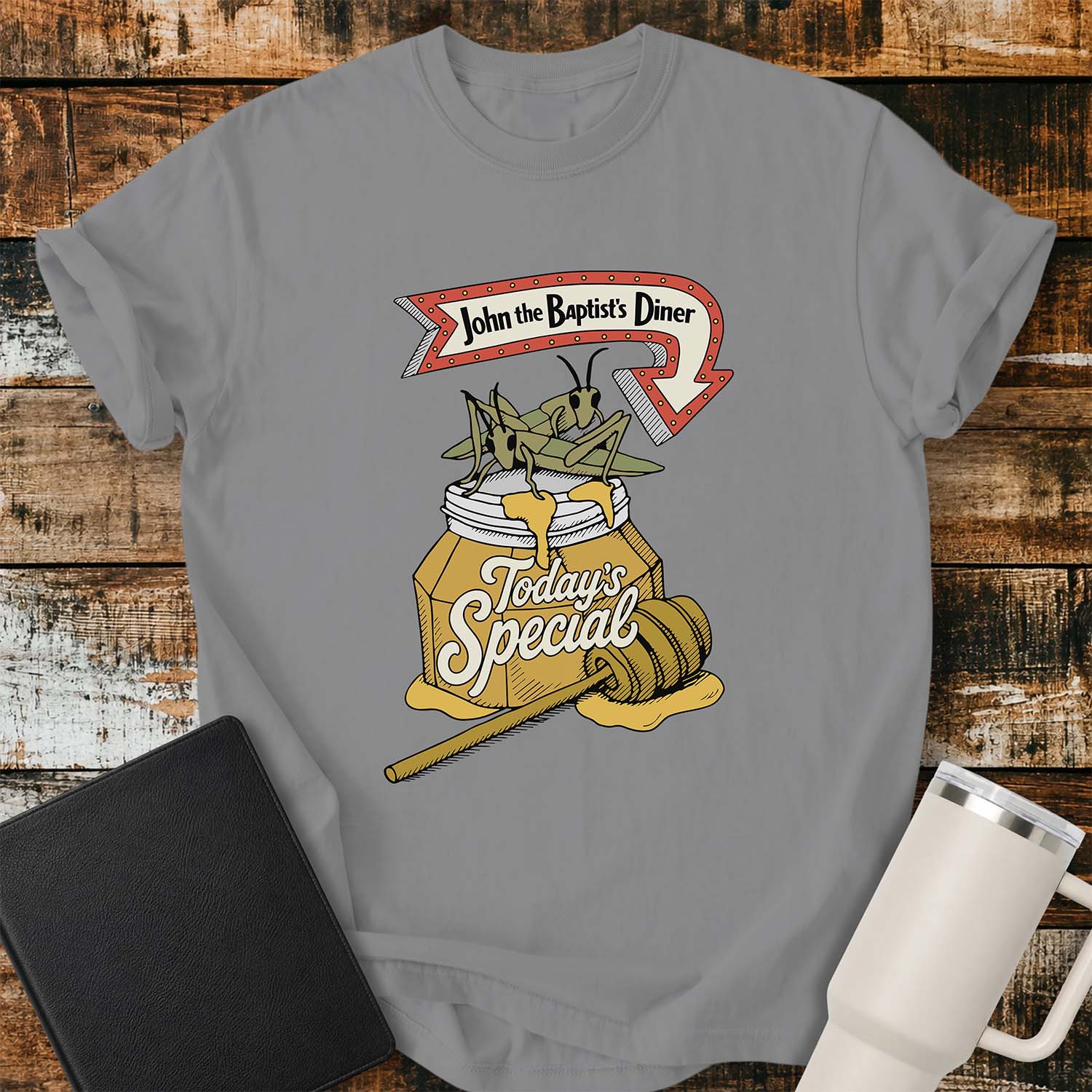 Gray T-shirt featuring a humorous vintage-inspired illustration for “John the Baptist’s Diner.” The design shows two large locusts perched on an overflowing jar of honey labeled “Today’s Special,” with a honey dipper resting beside it. A retro red arrow sign above reads “John the Baptist’s Diner.” The shirt is displayed on a rustic wood plank background with a black Bible on the left and a white insulated tumbler on the right.

