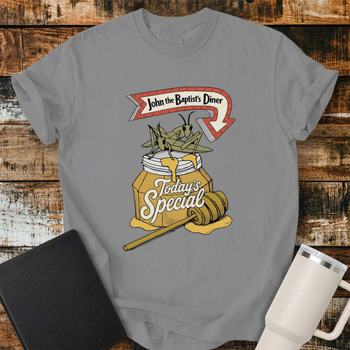 Gray T-shirt featuring a humorous vintage-inspired illustration for “John the Baptist’s Diner.” The design shows two large locusts perched on an overflowing jar of honey labeled “Today’s Special,” with a honey dipper resting beside it. A retro red arrow sign above reads “John the Baptist’s Diner.” The shirt is displayed on a rustic wood plank background with a black Bible on the left and a white insulated tumbler on the right.
