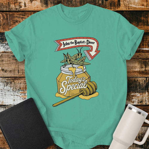 island green T-shirt featuring a humorous vintage-inspired illustration for “John the Baptist’s Diner.” The design shows two large locusts perched on an overflowing jar of honey labeled “Today’s Special,” with a honey dipper resting beside it. A retro red arrow sign above reads “John the Baptist’s Diner.” The shirt is displayed on a rustic wood plank background with a black Bible on the left and a white insulated tumbler on the right.
