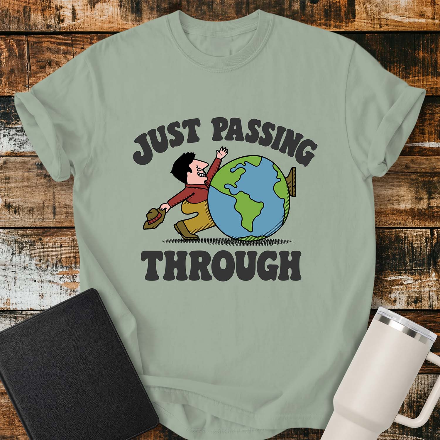 bay green T-shirt featuring a retro cartoon of a smiling traveler carrying a hat while walking behind a globe with the phrase “JUST PASSING THROUGH” in bold vintage lettering. The shirt is displayed on a rustic wood plank background alongside a black Bible and a white insulated tumbler.