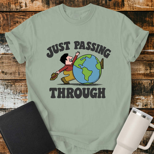 bay green T-shirt featuring a retro cartoon of a smiling traveler carrying a hat while walking behind a globe with the phrase “JUST PASSING THROUGH” in bold vintage lettering. The shirt is displayed on a rustic wood plank background alongside a black Bible and a white insulated tumbler.